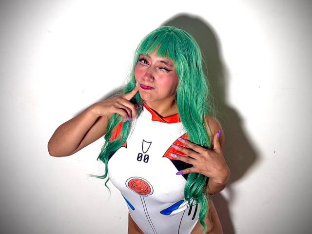 AilenRosse with green hair, wearing a white, red, and blue costume, touching her nose and chest, has purple nails, and a winking expression, standing against a white wall.