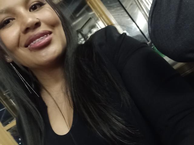 Novarose with long black hair, nose ring, black shirt, and pink lipstick, sticking out her tongue, showing her teeth, and wearing long earrings in a selfie.