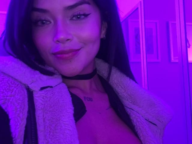 MajoJones con iluminación púrpura, sonriendo, collar negro, chaqueta blanca y esponjosa, sujetador negro, mostrando pechos pequeños, tatuaje sobre el pecho, cabello largo y oscuro, lápiz labial rojo, nariz ligeramente levantada..