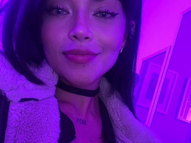 MajoJones, con el rostro iluminado en tonos morados, un collar negro, una chaqueta blanca y esponjosa y lápiz labial rojo, mostrando pequeños senos con un tatuaje que dice "POU" encima. Tiene el cabello largo y negro y una sonrisa sutil..