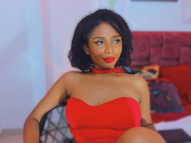 MeganWilliams with curly black hair, red lipstick, and a strapless red dress, showing her tits and a cute nose ring, sitting on a chair, smiling confidently.