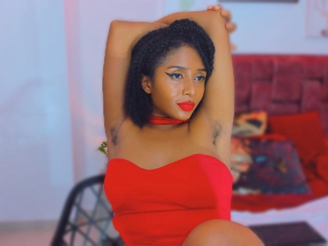 MeganWlliams with arms raised, showing armpit hair, wearing a red strapless dress, red choker, and red lipstick, looking at the camera with a slight smile.