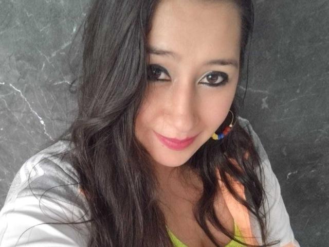 MelodyRosses has long dark hair, black eyeliner, pink lipstick, and a slight smile. She's wearing a white shirt and colorful earrings, with her tits slightly visible.