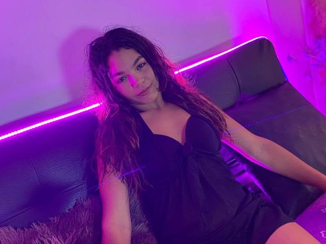 Susana69Foxy with wavy brown hair, black dress, and visible breasts, sitting on a black leather couch under purple neon lights. Her expression is relaxed and slightly seductive.