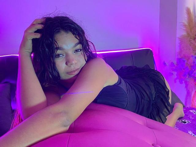 Susana69Foxy, con i capelli neri bagnati, è sdraiata su un divano rosa, indossa un vestito nero, guarda la fotocamera, con una luce al neon viola sullo sfondo, e ha una mano tra i capelli..