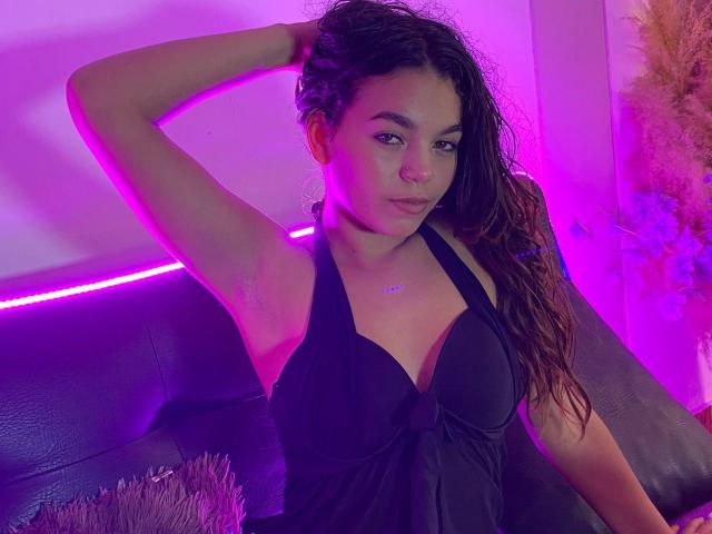 Susana69Foxy met nat bruin haar, een zwarte haltertopjurk, kleine borsten, arm omhoog, paars neonlicht, zittend op een zwarte leren bank, kijkend naar de camera met een lichte frons..