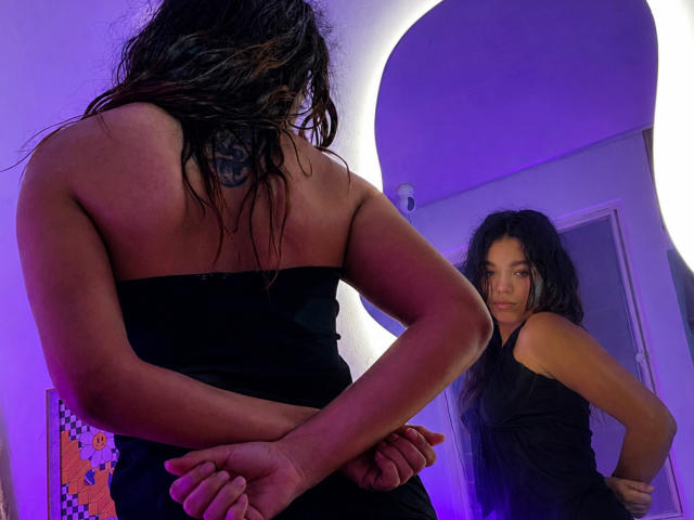 Susana69Foxy con capelli neri bagnati, tatuaggio sulla schiena, vestito nero senza spalline, braccia incrociate, che guarda il suo riflesso nello specchio, illuminazione viola, in piedi nella porta, mani sui fianchi..