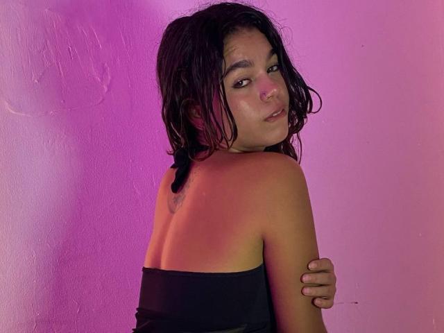 Susana69Foxy, avec des cheveux noirs et mouillés, un haut noir sans bretelles, regardant par-dessus son épaule, une main sur son bras, un mur rose-violet, un tatouage sur le haut du dos, un maquillage discret, une bouche légèrement ouverte, une expression confiante..