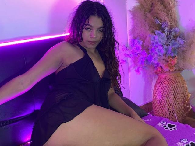 Susana69Foxy, avec de longs cheveux bruns bouclés, portant une robe noire transparente, montrant ses seins et ses cuisses pulpeuses, assise sur un canapé en cuir noir sous des lumières néon violettes..