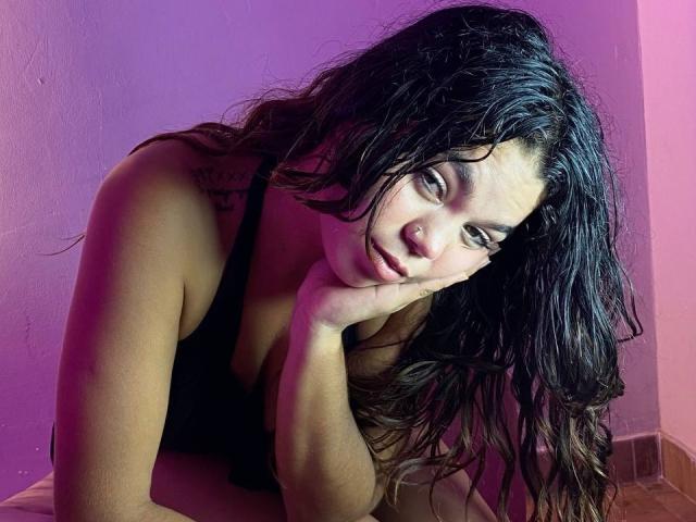 Susana69Foxy, avec ses cheveux noirs mouillés, appuyée sur sa main, portant un débardeur noir qui dévoile ses seins, éclairage violet et rose, et un tatouage sur son épaule gauche..