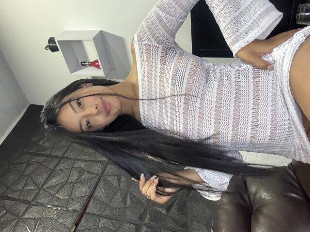 NicolethX, con cabello largo y negro, vestida con una blusa blanca de punto transparente, que deja ver sus pequeños senos firmes y su abdomen plano, está de pie con una mano en la cadera, sonriendo..