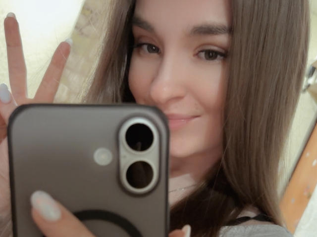 SmileForMee with long brown hair, holding a black phone in front of her face, smiling slightly, showing white nails, in a selfie. Her eyes are brown, and she's cute.