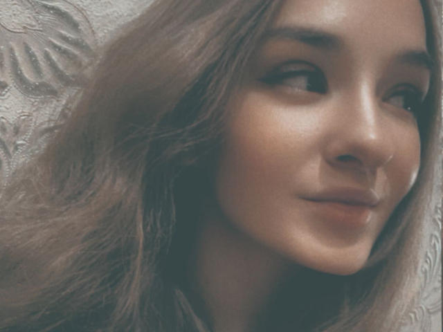 SmileForMee has wavy brown hair, a cute nose, and a subtle smile with soft makeup on her face, looking slightly to the side. Her skin is smooth and warm.