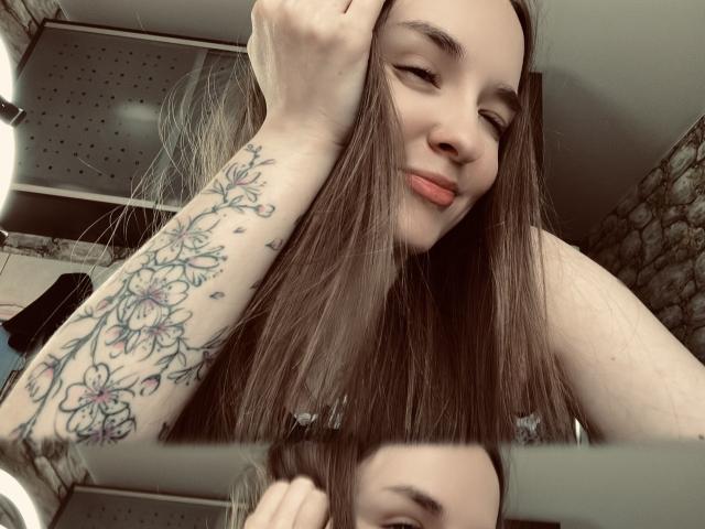 SmileForMee with long brown hair, holding her head, showing a tattooed arm with floral designs, smiling with red lips, in a mirror selfie.