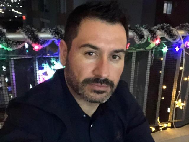 RodherX, un homme barbu avec des cheveux courts et foncés et une moustache, portant une chemise sombre, avec une expression sérieuse, dans un autoportrait avec des guirlandes de Noël colorées en arrière-plan..