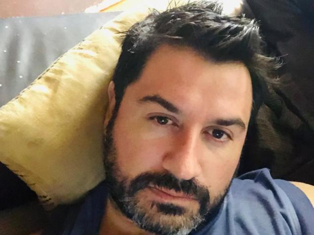 RodherX ha un viso paffuto, capelli scuri e disordinati, e una barba con un'espressione seria, sdraiato su un cuscino giallo con una camicia blu, che guarda direttamente verso la fotocamera..