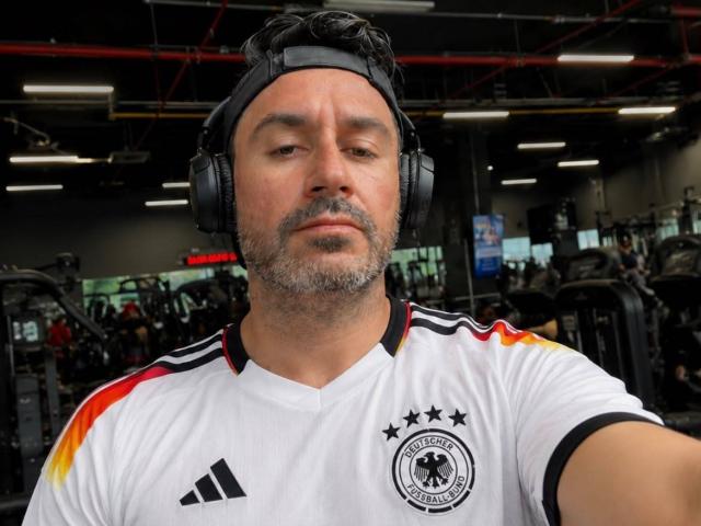 RodherX, un homme corpulent avec une barbe grise, portant un maillot de football allemand et des écouteurs noirs, a l'air sérieux dans une salle de sport. Son maillot a des rayures colorées sur les épaules..