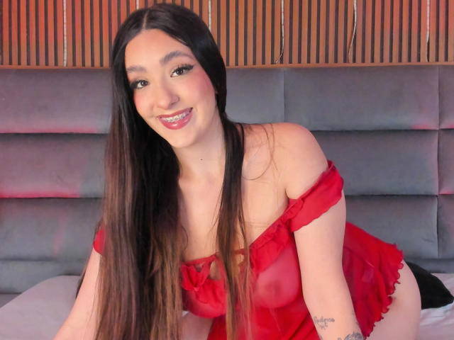 EliSanda, with long brown hair, red off-shoulder lingerie, and a smile, is kneeling on a bed, showing her tits and a tattoo on her right arm.