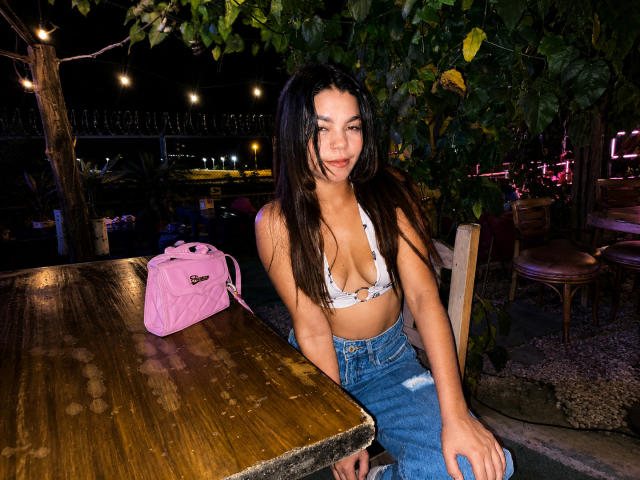 Susana69Foxy mit langen, dunklen Haaren, einem weißen BH-Top, das kleine Brüste zeigt, und blauen, hochgeschnittenen Jeans, sitzt an einem Holztisch mit einer pinken Tasche, Nacht, im Freien..