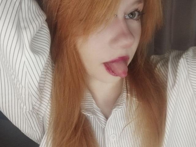 JamieLathrum has red hair, a striped shirt, and her tongue out, with her arm raised. She's wearing a small nose ring and has pink lipstick.
