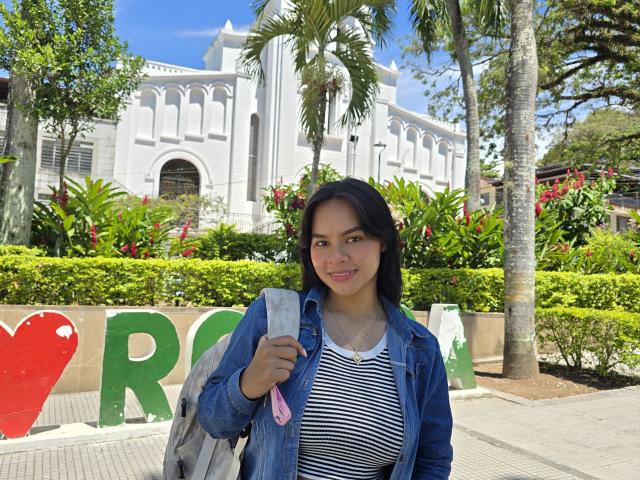 Yurimy, con i capelli scuri, seno piccolo, indossa una camicia a righe, una giacca di jeans e uno zaino grigio, ed è in piedi di fronte a un edificio bianco con palme e vegetazione..
