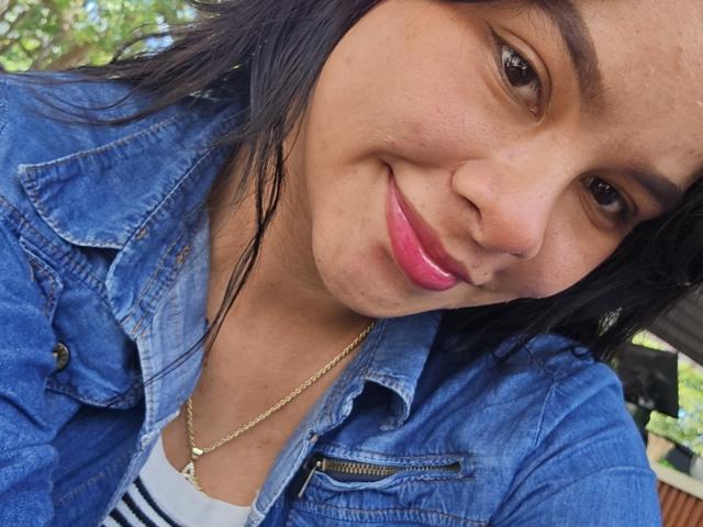 Yurimy's face, smiling with pink lipstick, dark eyes, and black hair, wearing a blue denim jacket and gold necklace, looking slightly to the right. Her tits are not visible.