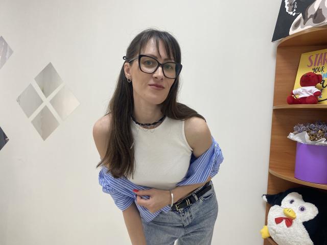MiraMixi portant des lunettes, avec un débardeur blanc, une chemise bleue à rayures portée par-dessus les épaules, un jean gris, un collier noir, et une main sur le ventre, debout dans une pièce avec une étagère et un pingouin en peluche..