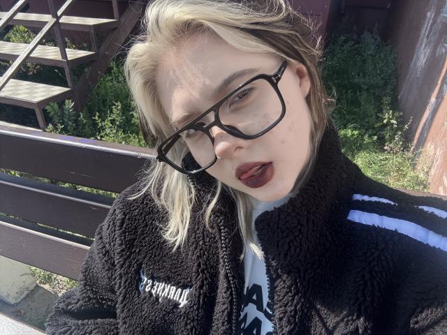 Krystal Berkovitch, con i capelli biondi, gli occhiali neri, il rossetto scuro e il giubbotto nero peloso, seduta all'aperto, guarda la fotocamera con la bocca leggermente aperta, mostrando un naso piccolo e la pelle pallida..