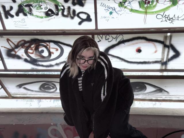 Krystal Berkovitch, mit blonden und schwarzen Haaren, die eine Brille trägt und einen schwarzen Pelzmantel anhat, hat einen ernsten Gesichtsausdruck und steht vor mit Graffiti beschmierten Fenstern, die Augenmuster zeigen..