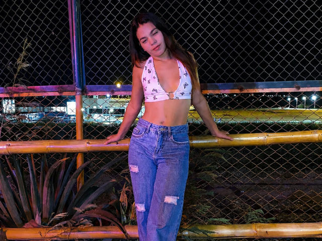 Susana69Foxy, con pechos pequeños, vestida con un top corto blanco de tirantes y jeans azules rotos, apoyada en una valla de bambú por la noche, mirando hacia abajo con una ligera sonrisa..