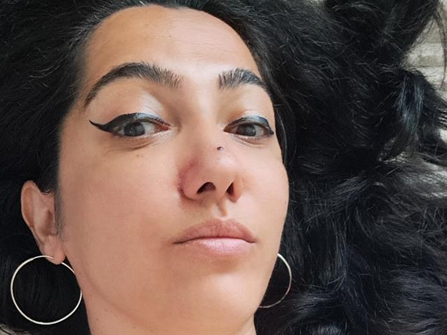SexyMirra's face with dark, wavy hair, bold black eyeliner, small nose piercing, pink lips, and large silver hoop earrings. Her skin is light brown, and she looks relaxed.