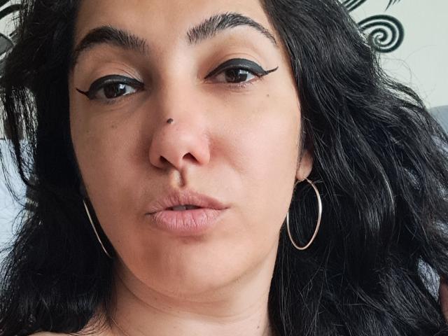 SexyMirra heeft donker, golvend haar, bruine ogen met zwarte, gevleugelde eyeliner, een kleine, zwarte beauty mark op haar neus, en ze frumelt met haar lippen, terwijl ze zilveren oorringen draagt..