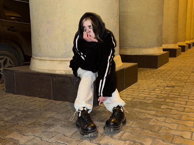 Krystal Berkovitch è accovacciata su un marciapiede in pietra, indossa una giacca da allenamento nera, pantaloni bianchi e stivali neri, con le mani vicino alla bocca, e sembra stanca o triste..