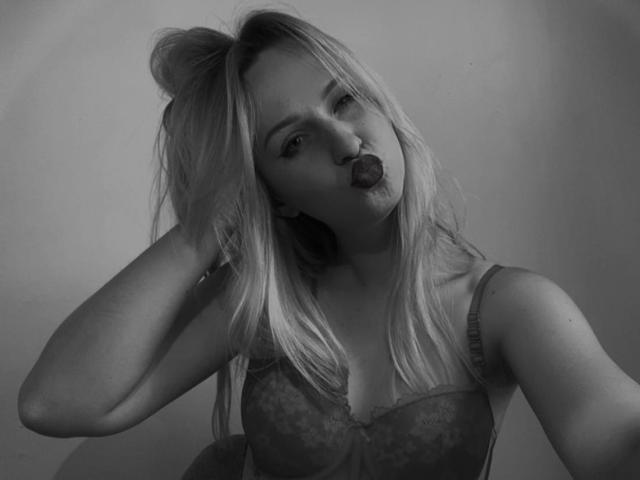FlyFreedom mit blonden Haaren, voller Lippen und herausgesteckter Zunge, trägt ein Spitzen-BH und berührt ihren Kopf, auf einem Schwarz-Weiß-Foto. Ihre Brüste sind sichtbar, mittelgroß..