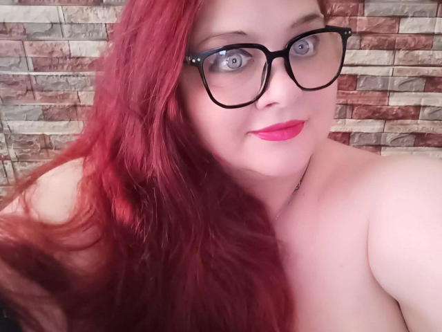 MolligeDani, con cabello rojo, gafas negras, ojos azules, lápiz labial rosa y hombros descubiertos, sonriendo a la cámara. No se ven los senos. En el fondo, una pared con un patrón de piedra..
