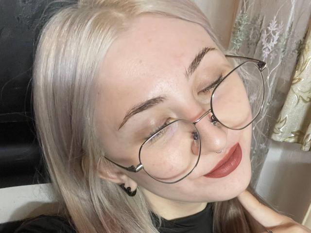 Carlene Agins, con cabello rubio, gafas, piercing en la nariz y lápiz labial rojo, sonriendo ligeramente, con los ojos cerrados, camisa negra y arete negro, tocándose el cuello..