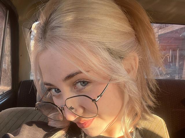 Carlene Agins, con una coleta rubia, gafas y labios fruncidos, con la luz del sol iluminando su rostro, sentada en un coche, vestida con una blusa negra, mirando a la cámara..