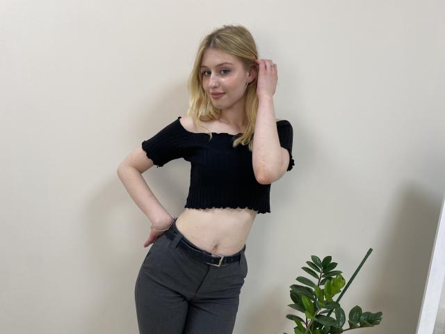 Nikollia, con cabello rubio, un top corto negro con un hombro descubierto y pantalones grises, de pie con una mano en la cadera, con un trasero pequeño y una cintura delgada, cerca de una planta verde..