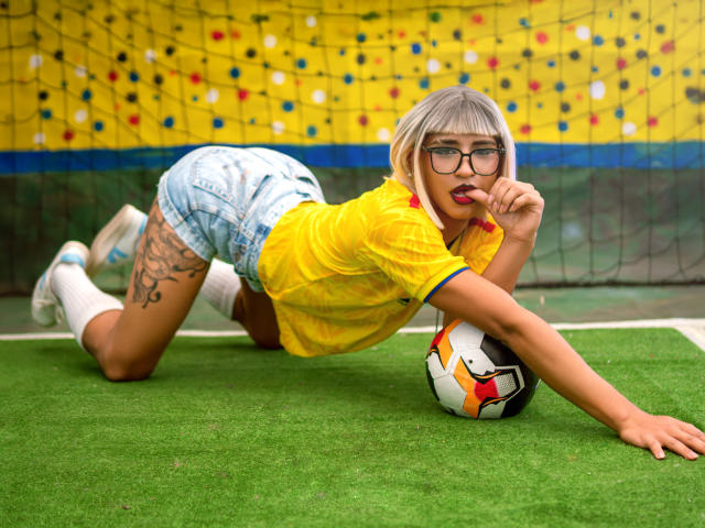 Kalyban, with blonde bob and glasses, kneels on green soccer field, wearing yellow shirt, blue plaid shorts, white socks, touching red lipstick, ball under her, tattoo on thigh.