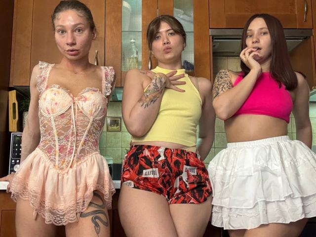 Drei Frauen von HotCaramelsTrio stehen in einer Küche, tragen Dessous und Sportbekleidung. Erste: Pinker Spitzenkorsett mit gerüschtem Rock, der die Brüste zeigt. Zweite: Gelbes Oberteil, rote Shorts mit Muster, die die Vulva zeigt. Dritte: Pinker Sport-BH, weißer gerüschter Rock, der das Gesäß zeigt..