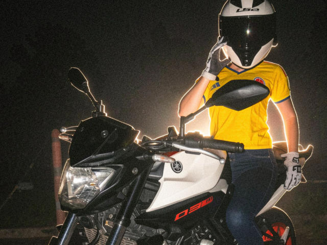 KimmyK with a black helmet, yellow shirt, and white gloves, sitting on a white and black Ducati motorcycle, wearing blue jeans, at night.