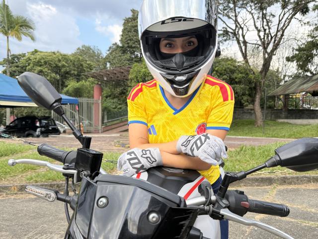 KimmyK wearing a shiny white helmet, yellow jersey with red and blue stripes, white gloves, and gray motorcycle, arms crossed, looking through helmet visor, black motorcycle in foreground.