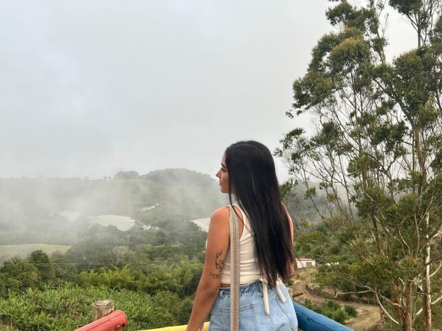 SharonKlart with long black hair, white tank top, and blue jeans, showing a tattoo on her right arm, standing on a foggy hill with trees and greenery.