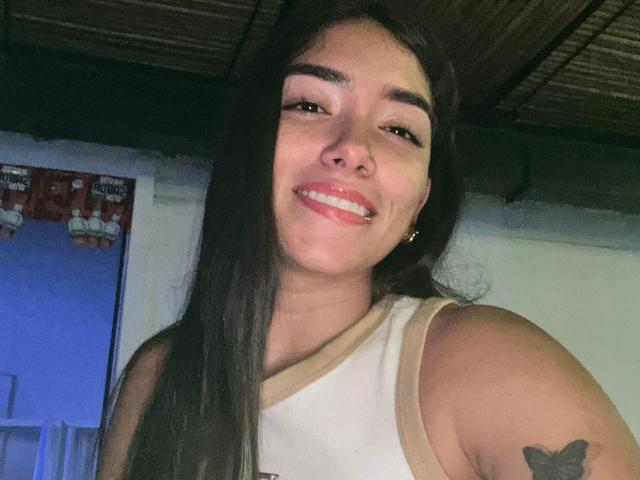 SharonKlart with long brown hair, smiling, wearing a white sleeveless top, showing a butterfly tattoo on her right arm, and small gold hoop earrings, in a dimly lit room.