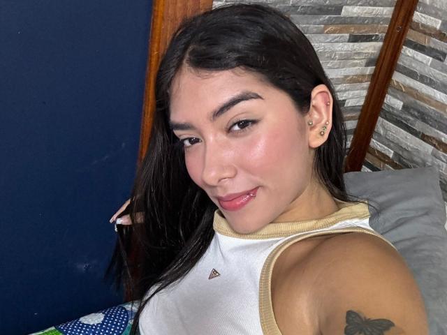 SharonKlart with dark hair, light skin, small nose, subtle makeup, pink lipstick, white sleeveless top, tattoo on upper arm, smiling, blue wall in background, stone-patterned wall.
