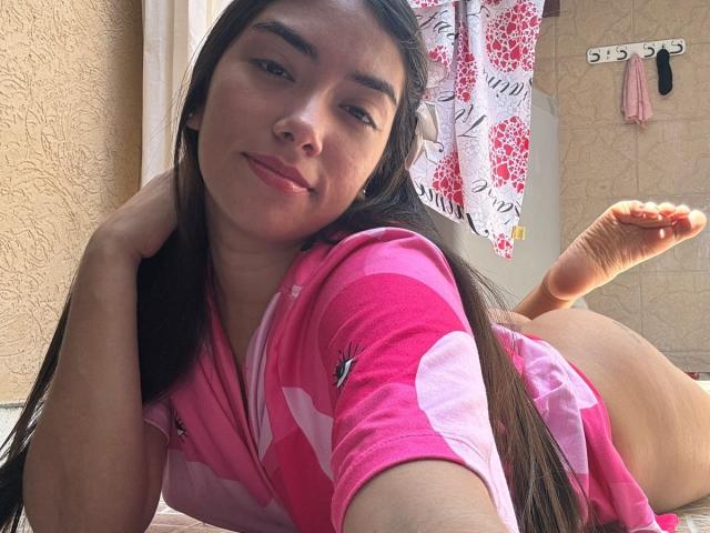 SharonKlart is lying on her stomach, wearing a pink shirt with white hearts, showing her bare ass. She has long brown hair, a slight smile, and is looking at the camera.