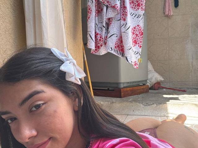 SharonKlart with dark hair, white bow, lying on floor, smiling, pink shirt, visible butt, small zit on cheek, laundry hanging, tiled floor, sunlight.