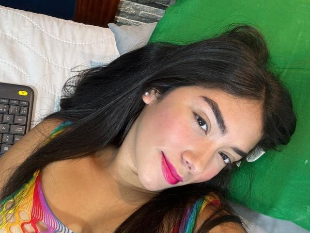 SharonKlart with long black hair, pink lipstick, and a colorful dress, lying on a green pillow, next to a calculator. Her tits are slightly visible under the dress.