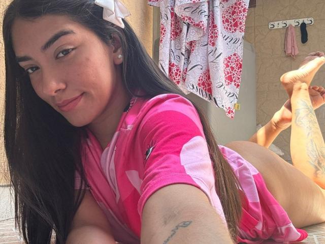 SharonKlart lying on her stomach, wearing a pink floral top, exposing her ass, with long black hair, white hairclip, tattoos on her arm and thigh, smiling at the camera.