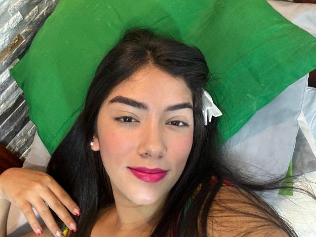 SharonKlart with long black hair, green pillow, pink lipstick, pearl earrings, dark eyebrows, and colorful nails, lying on bed, smiling slightly, holding pillow.